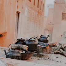 A series of black kettles are placed on outdoor stoves with logs of wood burning underneath them. The scene appears to be set in a rustic environment with earthen-colored walls and a cobblestone surface.
