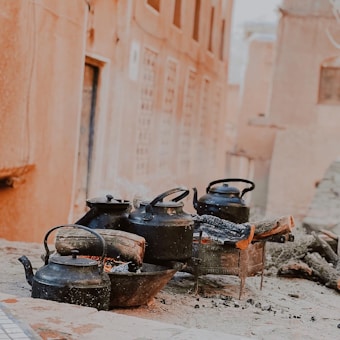 A series of black kettles are placed on outdoor stoves with logs of wood burning underneath them. The scene appears to be set in a rustic environment with earthen-colored walls and a cobblestone surface.