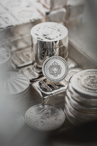 Stacks of shiny silver coins rest on top of silver bars, with intricate designs and engravings visible on the surfaces. The centerpiece coin features a lion’s head within a decorative border, while other coins display different motifs.