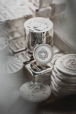 Stacks of shiny silver coins rest on top of silver bars, with intricate designs and engravings visible on the surfaces. The centerpiece coin features a lion&rsquo;s head within a decorative border, while other coins display different motifs.