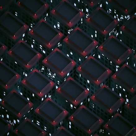 A geometric pattern of dark building-like structures arranged in a grid, each topped with a faintly glowing red light. The windows emit a soft bluish-white light, creating a contrast against the dark tones of the structures. The scene conveys a futuristic, urban atmosphere.