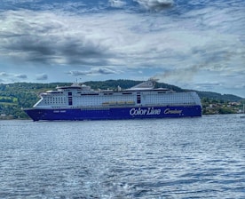 A large cruise ship with the words 'Color Line Cruises' on its side is sailing on a body of water. The ship is prominent with a blue and white color scheme, and visible lifeboats along the sides. In the background, a forested landscape and cloudy sky complete the scene.