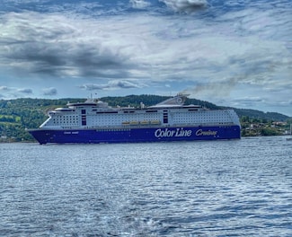 A large cruise ship with the words 'Color Line Cruises' on its side is sailing on a body of water. The ship is prominent with a blue and white color scheme, and visible lifeboats along the sides. In the background, a forested landscape and cloudy sky complete the scene.