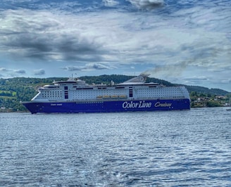 A large cruise ship with the words 'Color Line Cruises' on its side is sailing on a body of water. The ship is prominent with a blue and white color scheme, and visible lifeboats along the sides. In the background, a forested landscape and cloudy sky complete the scene.
