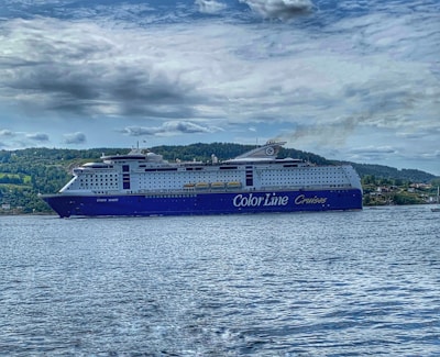 A large cruise ship with the words 'Color Line Cruises' on its side is sailing on a body of water. The ship is prominent with a blue and white color scheme, and visible lifeboats along the sides. In the background, a forested landscape and cloudy sky complete the scene.
