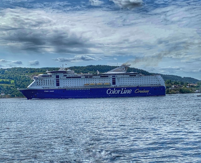 A large cruise ship with the words 'Color Line Cruises' on its side is sailing on a body of water. The ship is prominent with a blue and white color scheme, and visible lifeboats along the sides. In the background, a forested landscape and cloudy sky complete the scene.