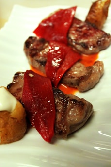 Grilled meat slices topped with red roasted peppers arranged on a white plate. A piece of bread with cream is visible on the side.