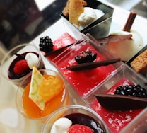 An assortment of colorful desserts served in small glasses, featuring vibrant red and orange mousses topped with various berries and decorative chocolate pieces. Whipped cream and small crumbs add texture to some of the servings, enhancing their visual appeal.