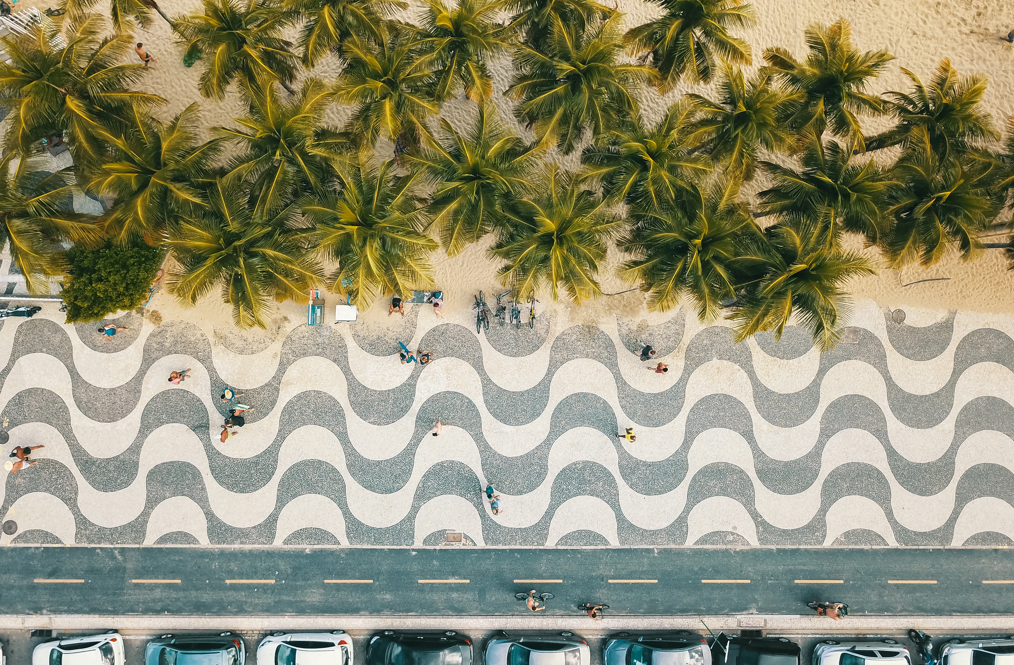 Overhead view of Copacabana's black-and-white wave-pattern mosaic promenade in Rio de Janeiro, a row of palm trees and the beach sand along one edge and the avenue along the other.