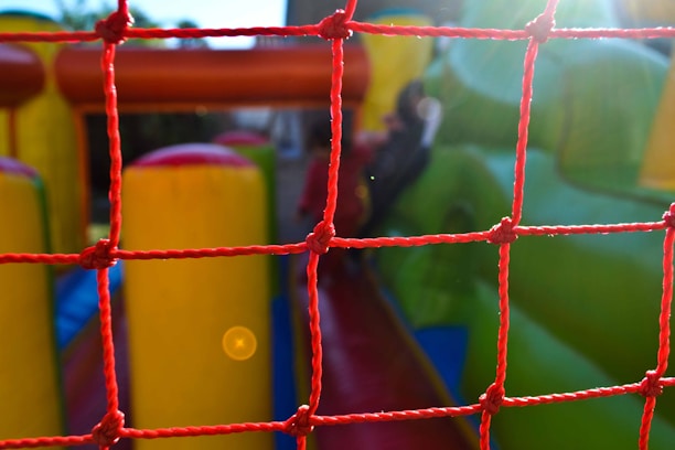 A red net is in the foreground, with a colorful inflatable play area visible behind it. The inflatable structure features bright green and yellow colors, hinting at a possible slide or obstacle course setup. Sunlight creates a lens flare, adding a warm glow to the scene.