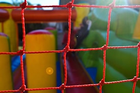 A red net is in the foreground, with a colorful inflatable play area visible behind it. The inflatable structure features bright green and yellow colors, hinting at a possible slide or obstacle course setup. Sunlight creates a lens flare, adding a warm glow to the scene.
