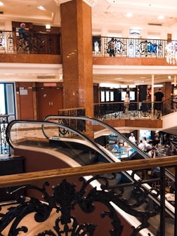 An elegant interior with two levels connected by an escalator. Ornate black iron railings with intricate designs decorate the space, combined with warm brown and cream tones. People are visible on both levels, engaging in conversations and enjoying the ambient atmosphere. A chandelier adds to the opulent setting, and natural light filters through large windows.