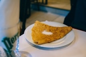 A folded crepe placed on a white plate with a small amount of sugar sprinkled on top. The setting appears to be a casual dining environment with soft lighting. A glass with a green logo and a napkin can be seen blurred in the foreground.