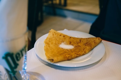 A folded crepe placed on a white plate with a small amount of sugar sprinkled on top. The setting appears to be a casual dining environment with soft lighting. A glass with a green logo and a napkin can be seen blurred in the foreground.