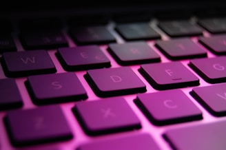 Close-up view of a computer keyboard with a vibrant pink and purple lighting effect. The keys are slightly angled, and the letters are clearly visible, giving a modern and tech-oriented vibe.