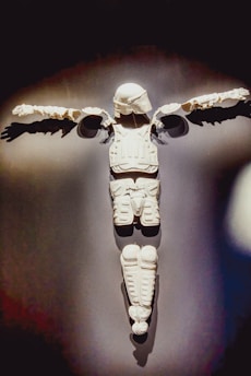 White body armor arranged on a dark background to resemble the shape of a person with arms outstretched, composed of various padded sections including a helmet.