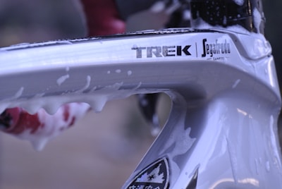 Close-up of a bicycle frame with a shiny silver finish, featuring brand logos such as TREK and Segafredo. The frame appears to be covered in soap suds, suggesting it is being cleaned. A hint of a red and black object is visible in the background.