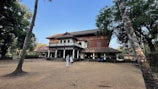 A large, traditional-style building with a sloped roof is surrounded by trees and an open sandy area. Several people in traditional white attire are gathered near the entrance of the building, suggesting a cultural or historical setting.