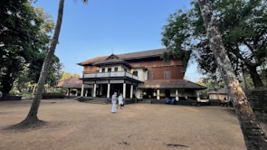 A large, traditional-style building with a sloped roof is surrounded by trees and an open sandy area. Several people in traditional white attire are gathered near the entrance of the building, suggesting a cultural or historical setting.