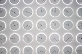 A wall is covered with multiple clocks arranged in a grid pattern. Each clock shows a different time and is encased in a circular frame with minimalistic black hands against a white background. The clocks are evenly spaced, creating a uniform and organized appearance.