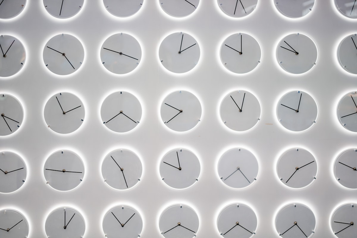 35 simple grey clocks set to various times on a grey wall.  Each clock has a white glow