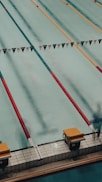 A swimming pool with several lanes marked by colorful lane dividers, including red and gold. Starting blocks are visible at the pool's edge.