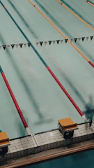 A swimming pool with several lanes marked by colorful lane dividers, including red and gold. Starting blocks are visible at the pool's edge.