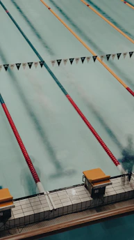 A swimming pool with several lanes marked by colorful lane dividers, including red and gold. Starting blocks are visible at the pool's edge.