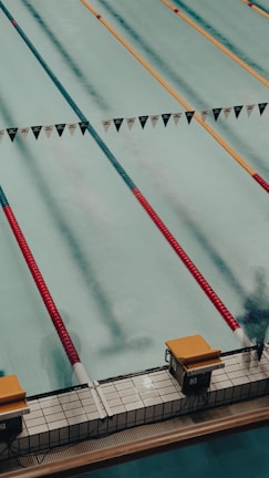 A swimming pool with several lanes marked by colorful lane dividers, including red and gold. Starting blocks are visible at the pool's edge.