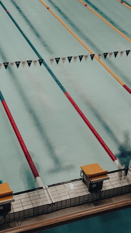 A swimming pool with several lanes marked by colorful lane dividers, including red and gold. Starting blocks are visible at the pool's edge.