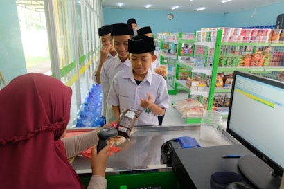 A group of boys in uniform stand in line at a convenience store checkout counter. They are wearing black caps and white shirts, and the cashier is scanning an item with a barcode scanner. The store shelves are stocked with various products, including snacks and bottled water. A computer screen is visible on the counter.