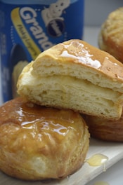 Golden, flaky biscuits drizzled with honey sit on a light wooden surface, with a can of biscuit dough visible in the background.