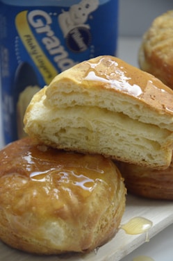 Golden, flaky biscuits drizzled with honey sit on a light wooden surface, with a can of biscuit dough visible in the background.