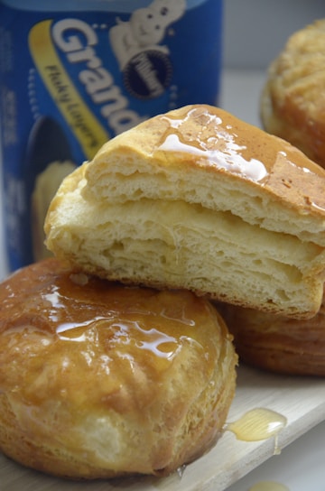 Golden, flaky biscuits drizzled with honey sit on a light wooden surface, with a can of biscuit dough visible in the background.