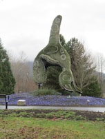 A large topiary sculpture designed in the shape of an orca whale is the central focus, surrounded by well-manicured bushes and a ground covering of small purple-blue flowers. The setting is an outdoor park with a bench on the left and various trees in the background, against a cloudy sky.