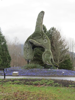 A large topiary sculpture designed in the shape of an orca whale is the central focus, surrounded by well-manicured bushes and a ground covering of small purple-blue flowers. The setting is an outdoor park with a bench on the left and various trees in the background, against a cloudy sky.