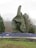 A large topiary sculpture designed in the shape of an orca whale is the central focus, surrounded by well-manicured bushes and a ground covering of small purple-blue flowers. The setting is an outdoor park with a bench on the left and various trees in the background, against a cloudy sky.
