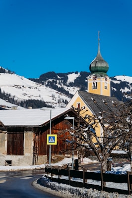 A picturesque winter scene featuring a snow-covered landscape with a traditional European church in the foreground. The church has a distinctive green onion dome and a yellow facade. Snow-laden roofs and leafless trees line a narrow road, with a pedestrian crossing sign visible. In the background, snow-capped mountains and evergreen forests are illuminated by bright sunlight under a clear blue sky.