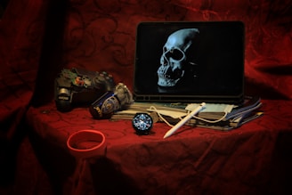 A dark, moody scene features a tablet displaying an image of a skull. Beside it, a worn game controller lies next to a crumpled NYPD can. A stack of documents is partially covered by a white stylus. A face mask and a round digital clock or watch are also visible. The setting rests on a deep red, intricately patterned fabric.