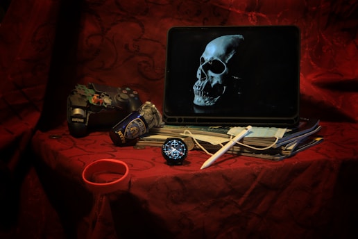A dark, moody scene features a tablet displaying an image of a skull. Beside it, a worn game controller lies next to a crumpled NYPD can. A stack of documents is partially covered by a white stylus. A face mask and a round digital clock or watch are also visible. The setting rests on a deep red, intricately patterned fabric.