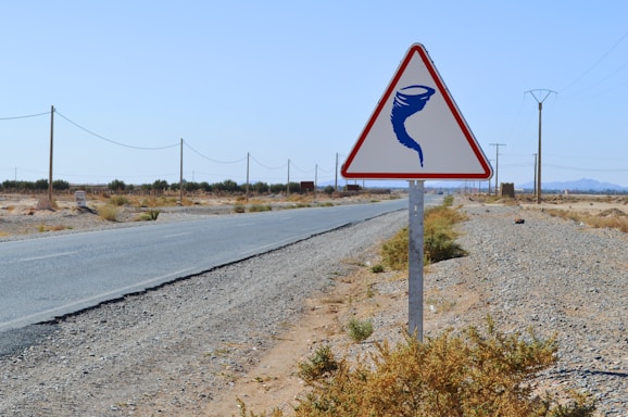 A triangular warning sign with a blue tornado symbol is placed beside a deserted road. The landscape is arid and flat, with sparse vegetation and utility poles lining the road. The sky is clear and the distant mountains create a horizon.