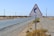 A triangular warning sign with a blue tornado symbol is placed beside a deserted road. The landscape is arid and flat, with sparse vegetation and utility poles lining the road. The sky is clear and the distant mountains create a horizon.
