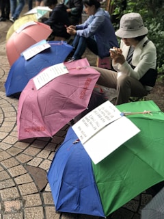Several colorful umbrellas with attached signs are placed on the ground in an outdoor setting. A person wearing a hat and casual clothing is seated nearby, looking at a phone. Other individuals are sitting in the background, possibly engaged in an outdoor activity or gathering.