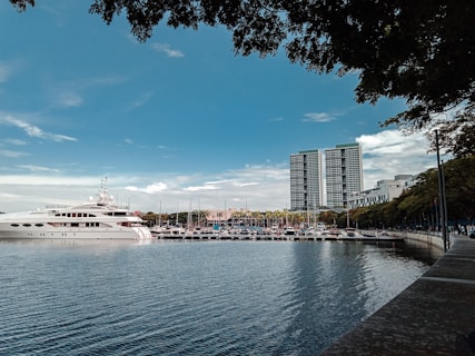 A large yacht is docked in a marina with several smaller boats and a cityscape in the background. Two tall buildings stand prominently against a partly cloudy sky. The foreground features calm water reflecting the scene, with lush green trees framing the top and right edges.