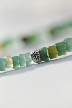 A close-up view of a string of small, round beads in shades of green, with one silver bead featuring an engraved leaf pattern. The beads appear to be made of glass or a similar translucent material, creating a natural and artisanal look.