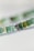 A close-up view of a string of small, round beads in shades of green, with one silver bead featuring an engraved leaf pattern. The beads appear to be made of glass or a similar translucent material, creating a natural and artisanal look.