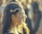 A woman with braided hair adorned with a decorative headpiece is seen in profile. She is wearing long earrings, and the sunlight creates a glowing effect on her hair. The background is blurred, suggesting a crowd or an outdoor gathering.