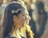 A woman with braided hair adorned with a decorative headpiece is seen in profile. She is wearing long earrings, and the sunlight creates a glowing effect on her hair. The background is blurred, suggesting a crowd or an outdoor gathering.