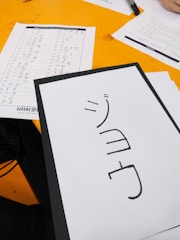 A Japanese katakana worksheet is on a yellow desk. In the foreground is a sheet with katakana characters, next to a completed practice sheet with columns of kana, possibly from a language learning session.