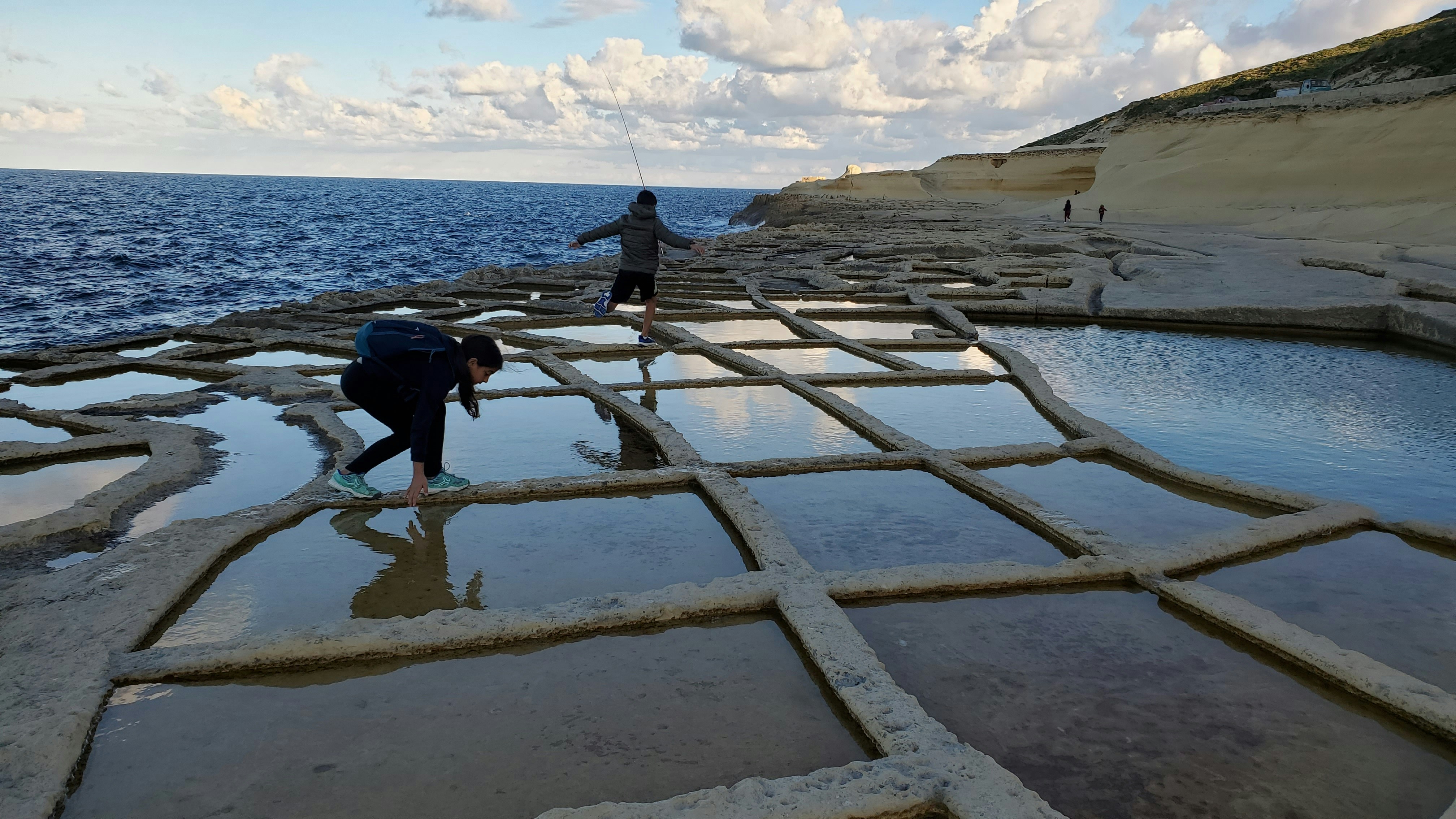 two people standing on a rocky beach next to the ocean, Salt Pans in Gozo, Malta 